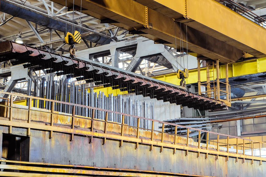 Two pulleys on a crane ready to lower a steel grid with rows of rods hanging to be dipped into molten zinc