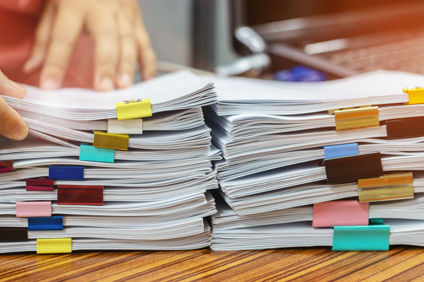 Papers representing cycling research are held together by a rainbow of binder clips are stacked on a desk