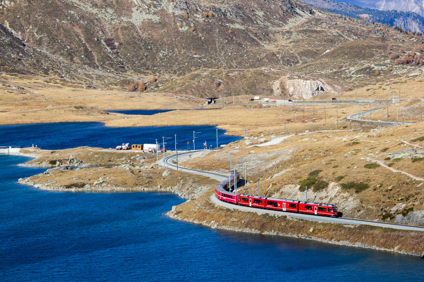 A red train snakes through a river valley in Switzerland